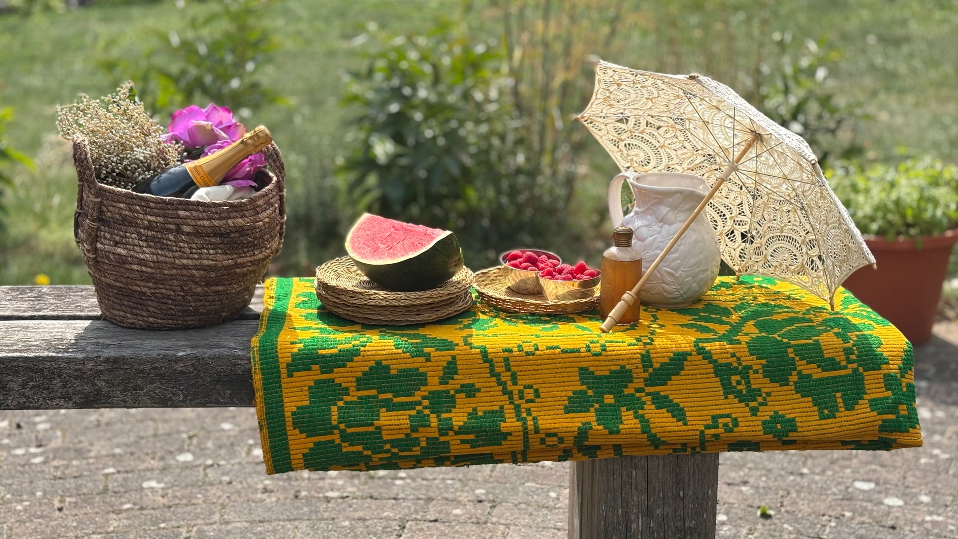 Outdoor setting with a colorful tablecloth, watermelon, and decorative items on a bench.