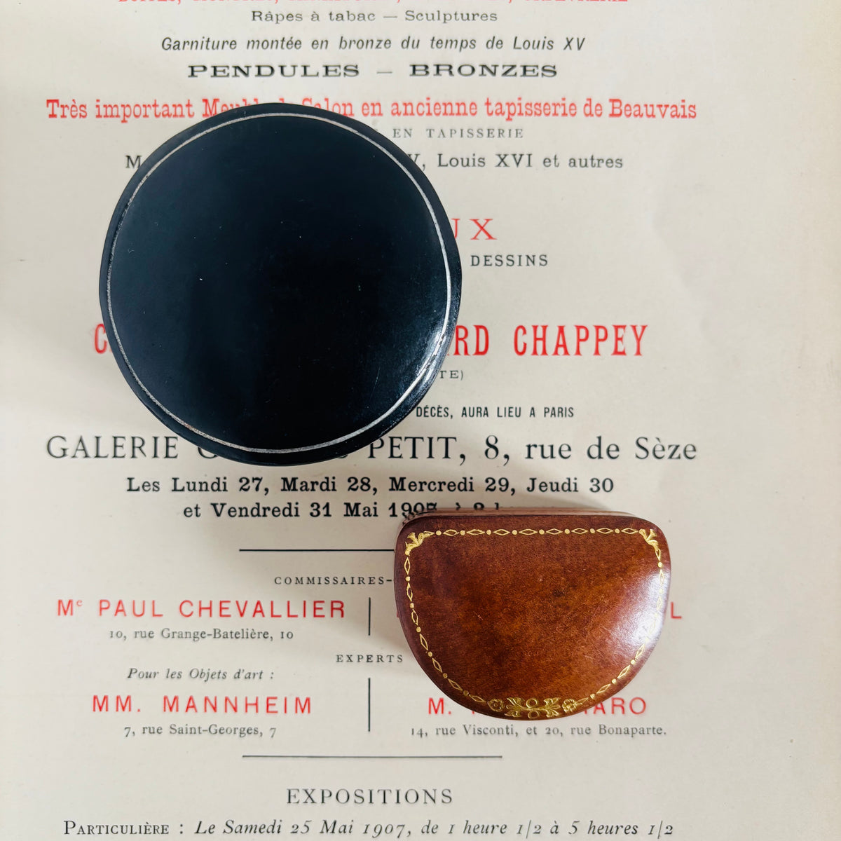 A vintage leather case with gold trim and white stitching, shown next to a round black object on top of a printed vintage poster.