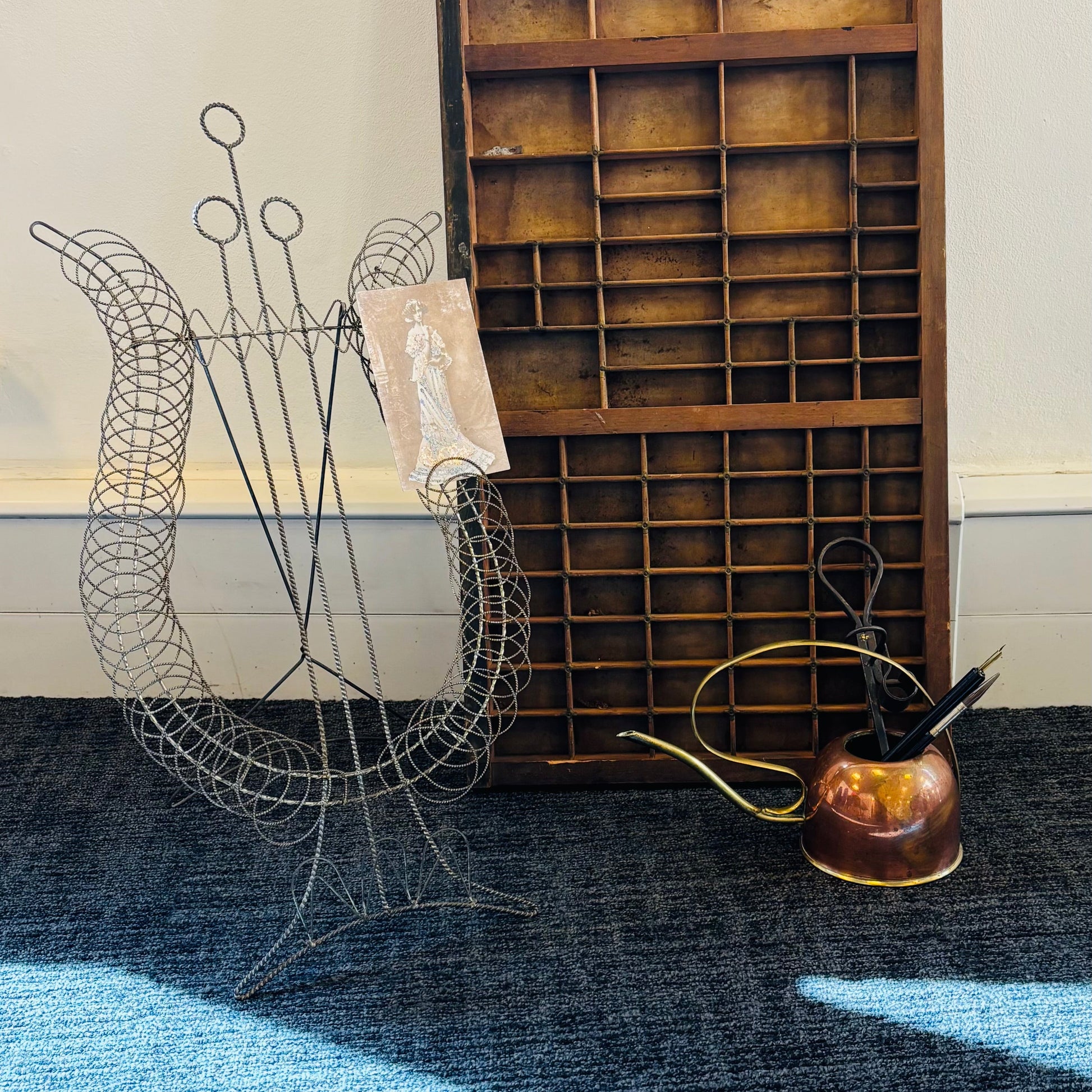 A tall wooden letterpress printing block board with multiple compartments, standing on a dark blue carpet next to a wire chair stand and a small copper pot.