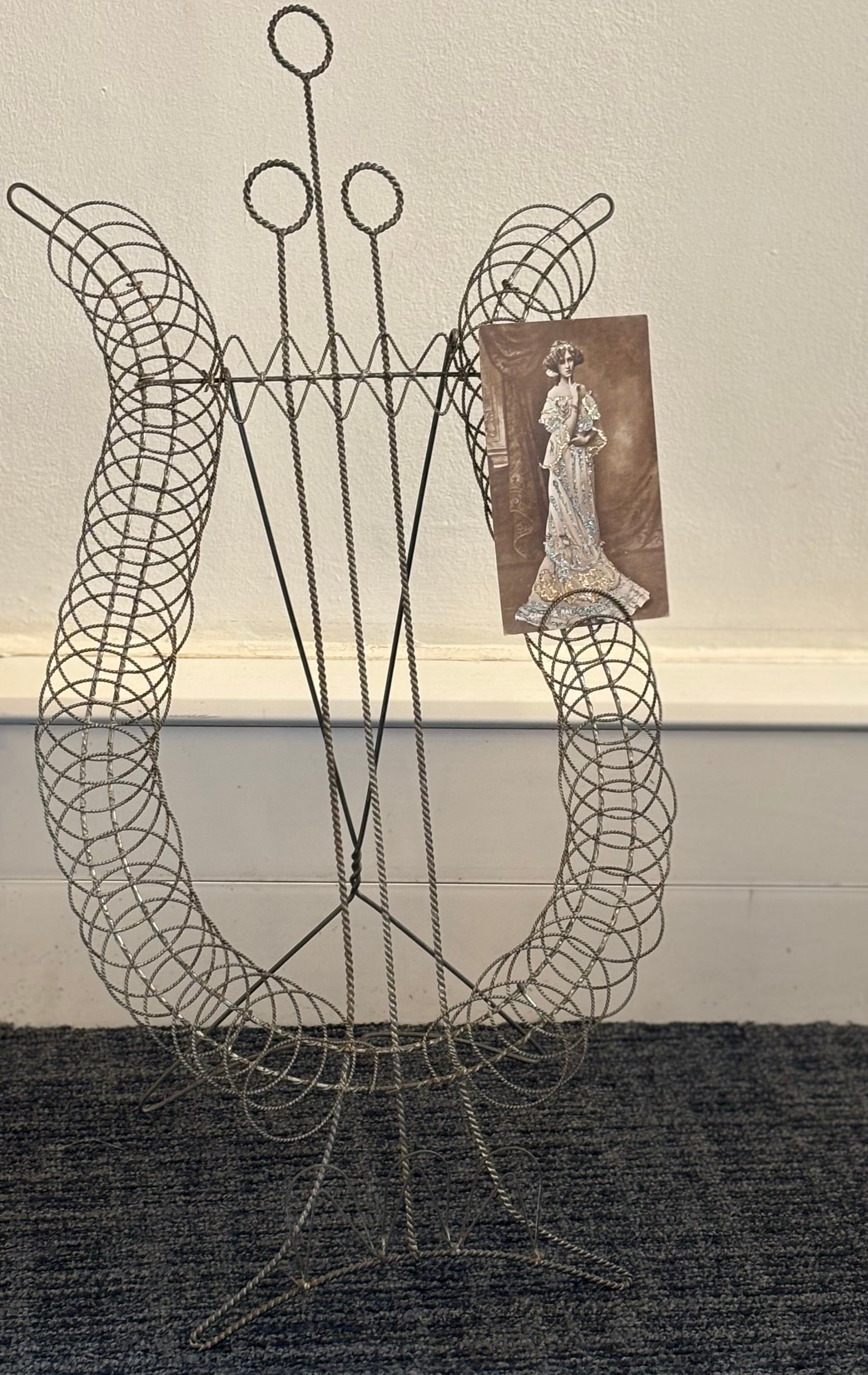 A vintage-style metal wire display stand with three upright loops and a curved base, shown standing on a dark textured rug against a white wall with a baseboard. A small brown vintage card depicting a woman in a gown is attached to the right side of the stand.