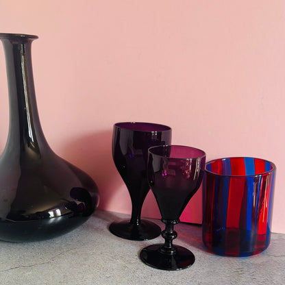 Amethyst wine glassware and a red and blue striped glass against a pink background