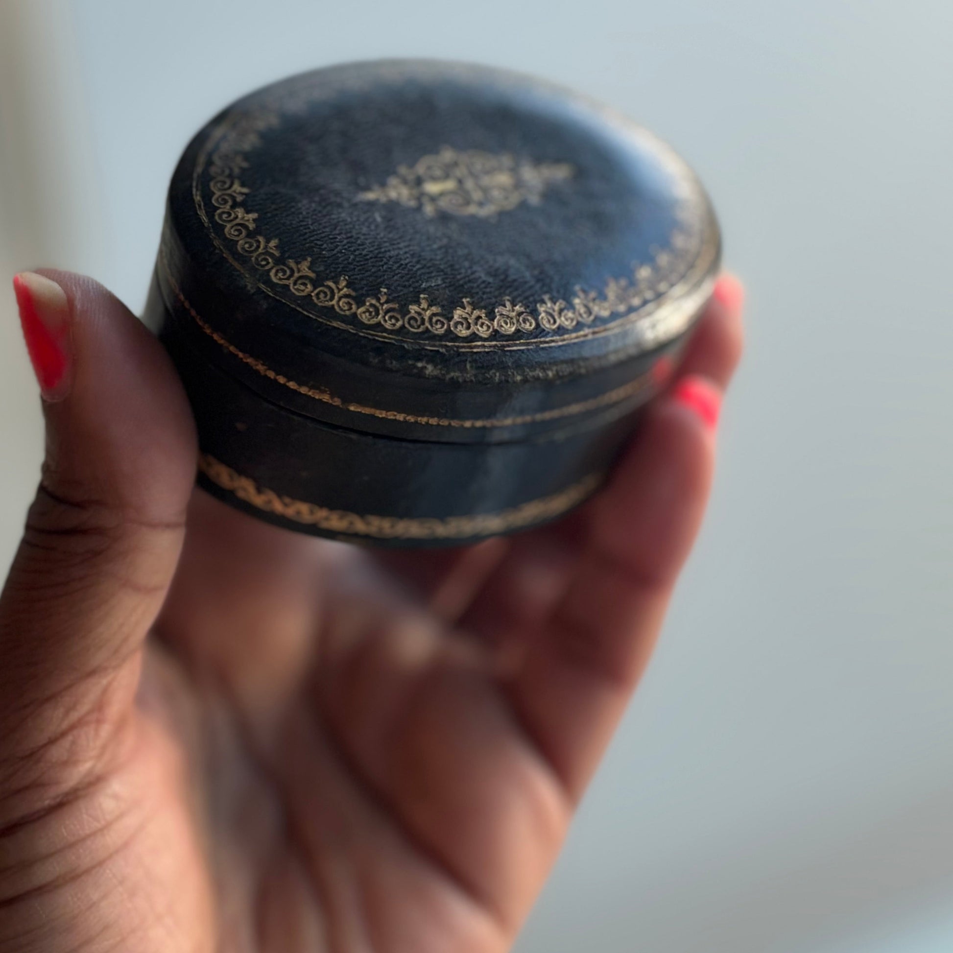 Hand holding a small round black container with gold detailing against a light background