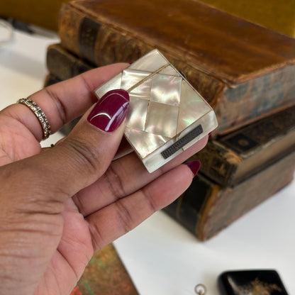 Hand holding a small mother of pearl vesta  on a white surface with books 