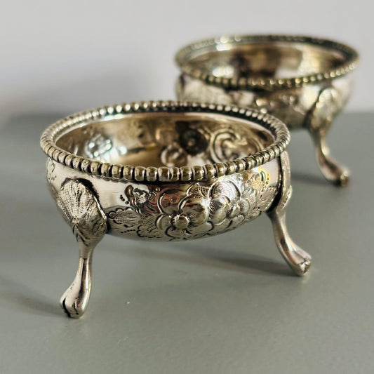 Two ornate silver salts with intricate designs on a gray surface.