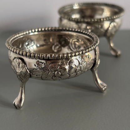 Two ornate silver salt cellers with intricate designs on a grey surface.