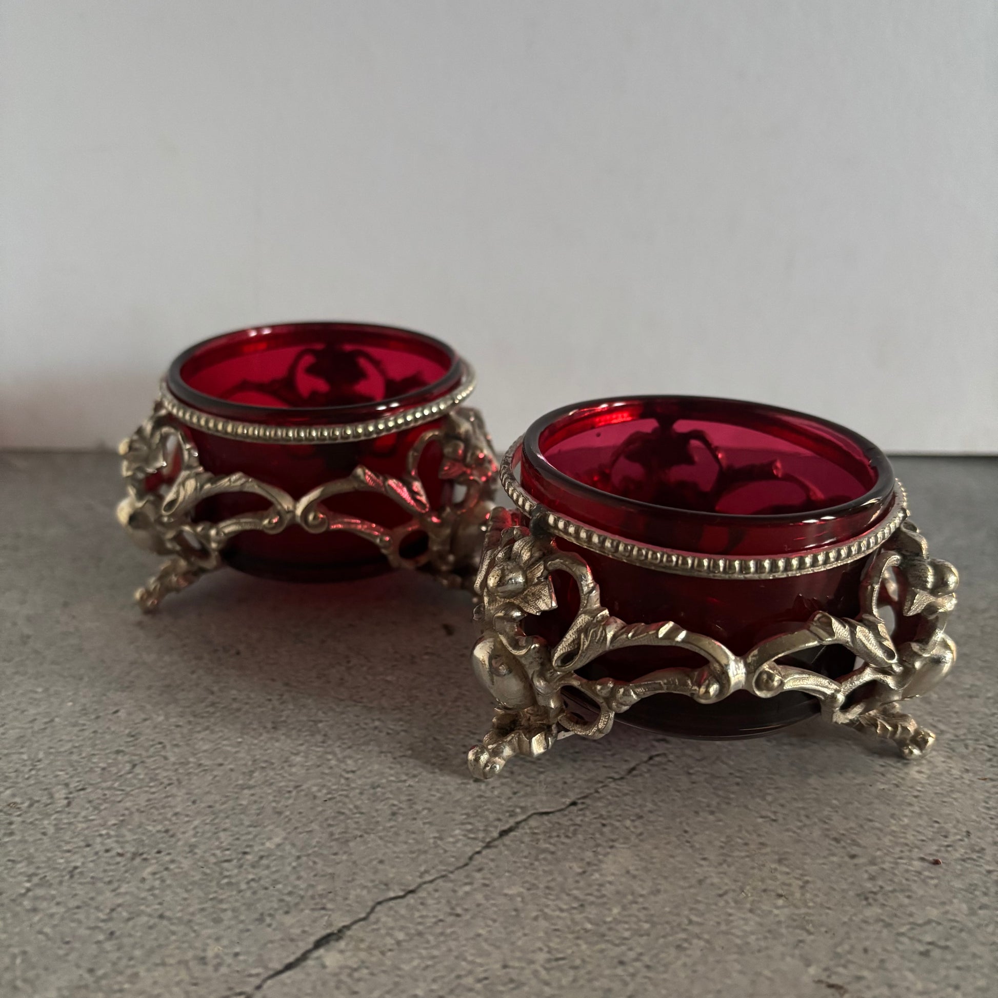 Two ornate red glass containers with silver metallic bases on a grey surface.