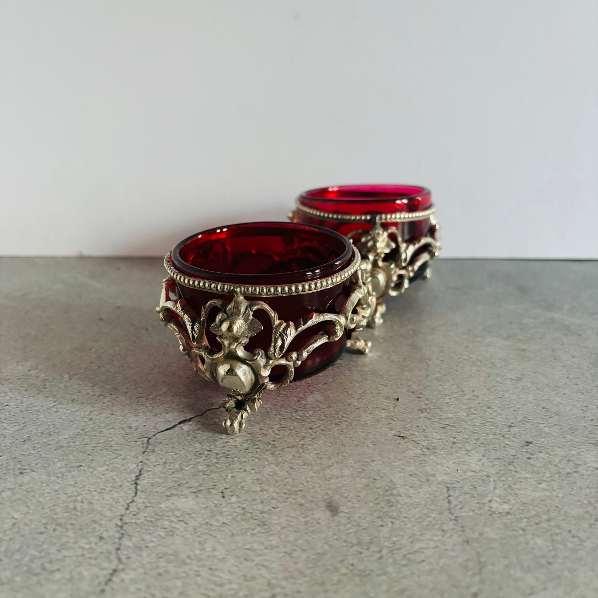 Two ornate glass containers with red glass and silver metal bases on a gray surface.