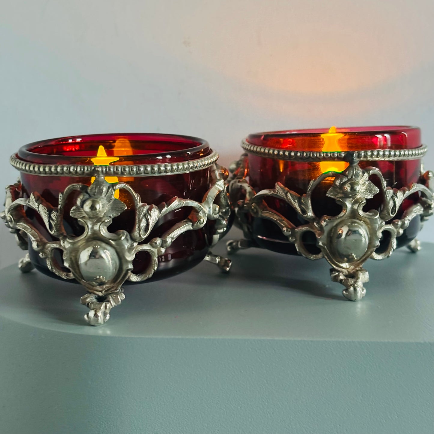 Two ornate candle holders with red glass and antique silver on a grey surface.