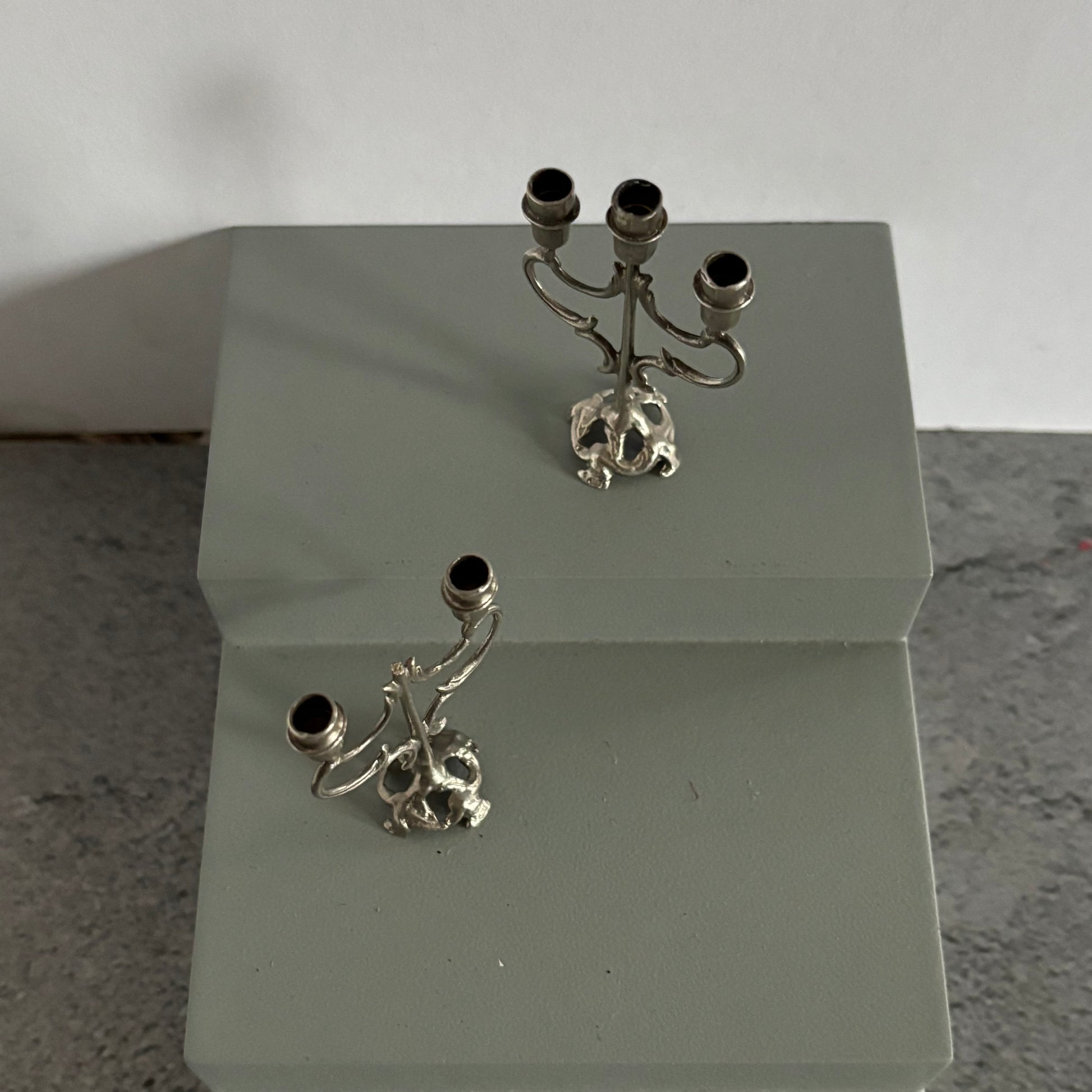 Two silver candelabra with skull designs on a white stand against a light gray background.