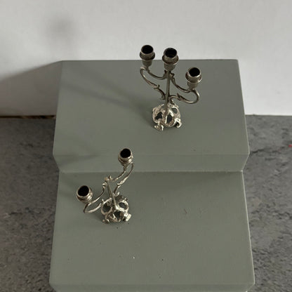 Two silver candelabra with skull designs on a white stand against a light gray background.