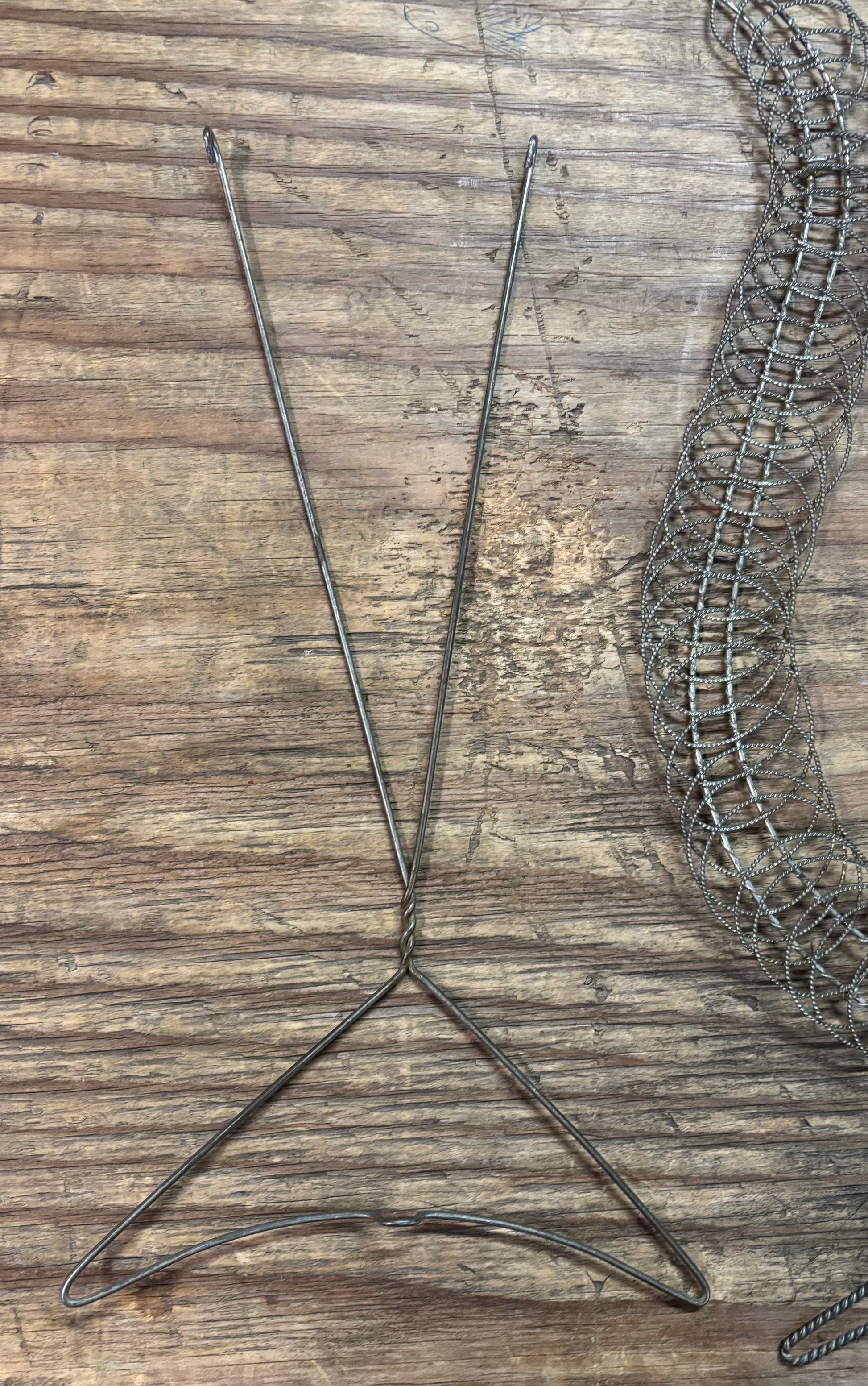 A metal wire hanger stand with three long legs, shown on a rustic wooden plank surface next to a coiled wire mesh.