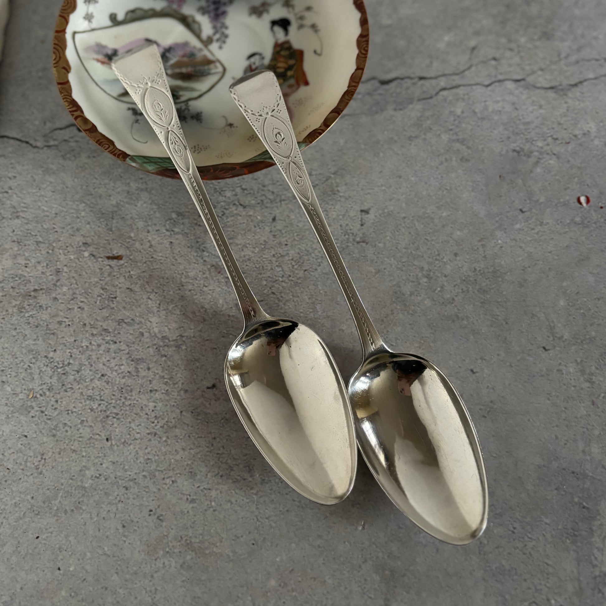 Two silver spoons on a textured gray surface with a decorative dish in the background.