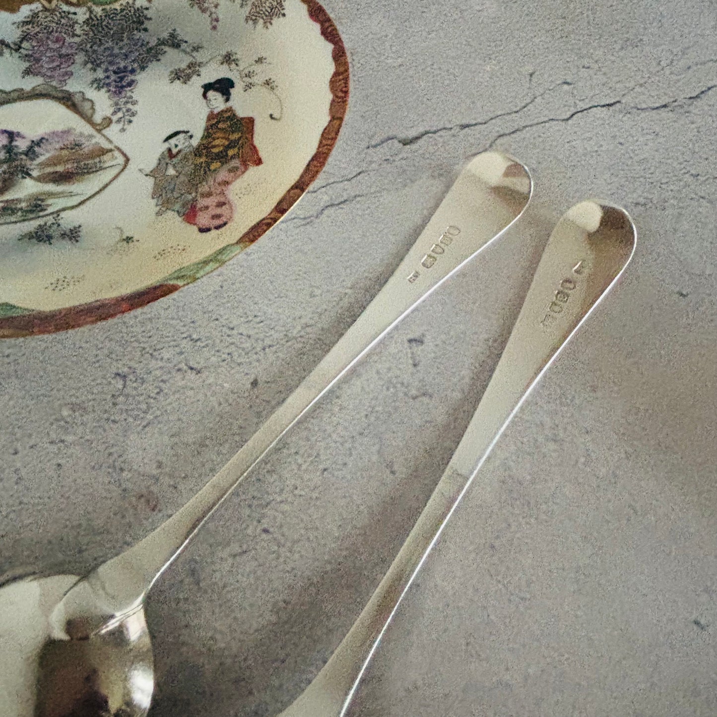 Two silver spoons on a textured surface with a decorative plate in the background.