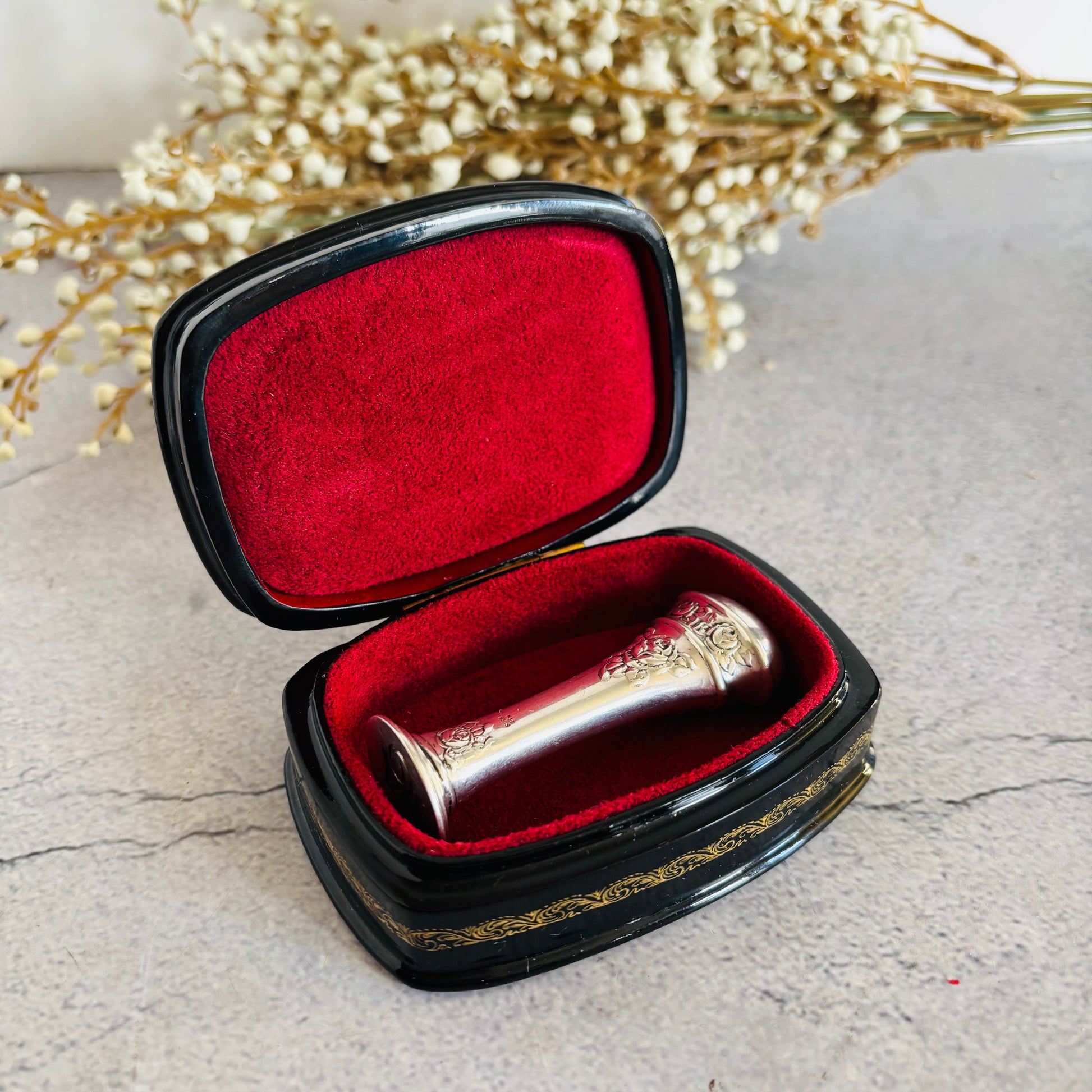 Silver wax seal stamp  in an open black case with red interior, placed on a textured surface with dried flowers.