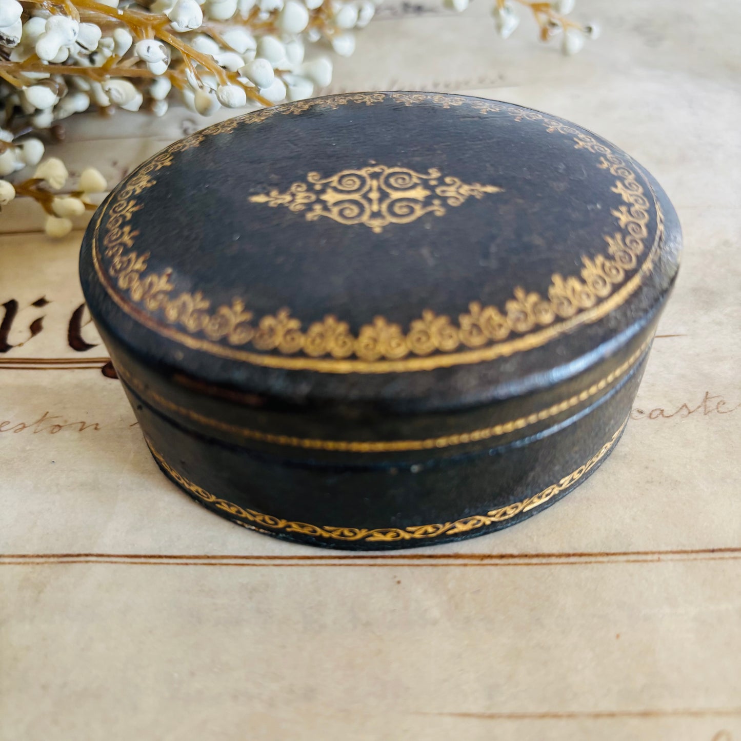 Round black box with gold detailing with white flowers in the background