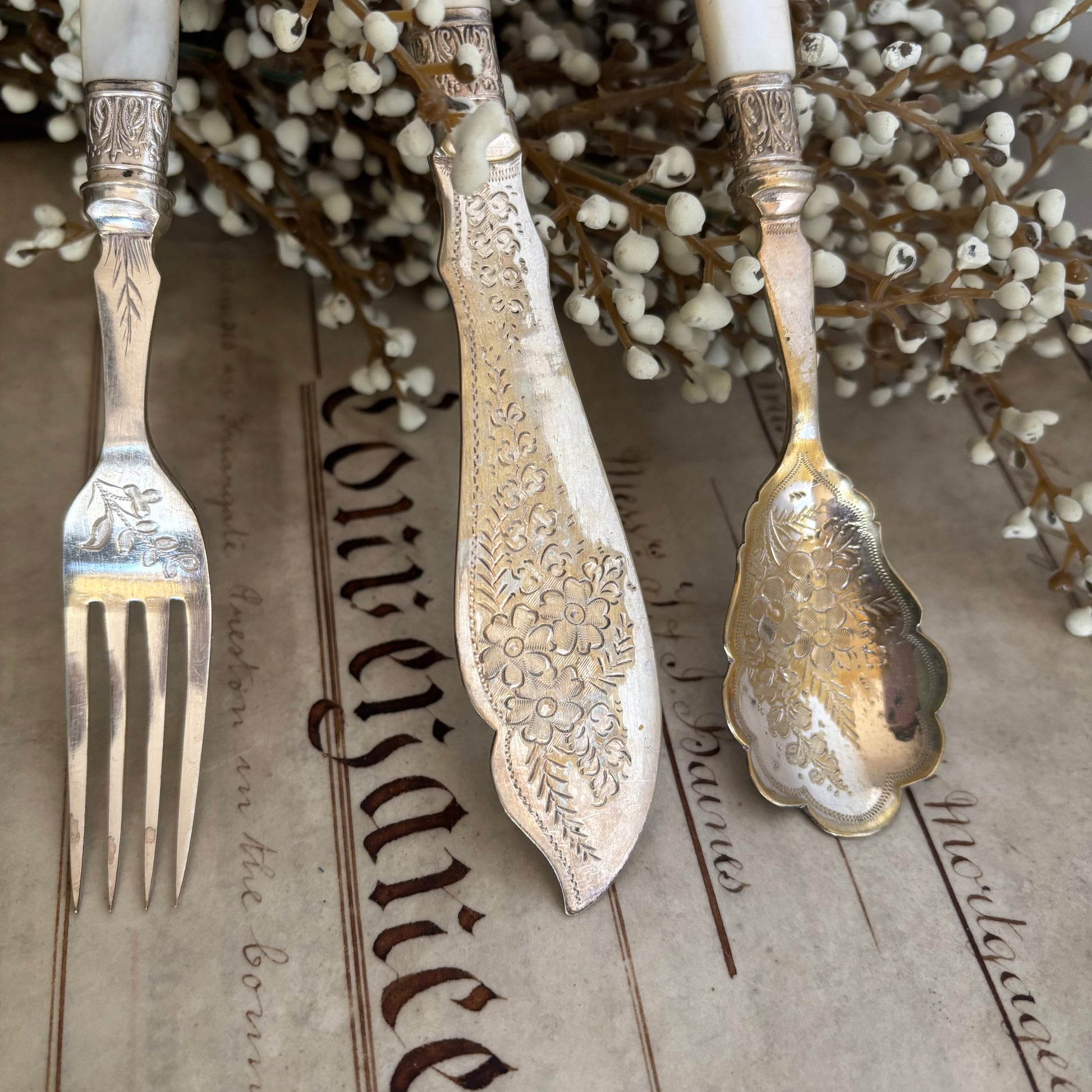Decorative silver fork, spoon, and knife with intricate patterns on a textured surface with white flowers in the background.