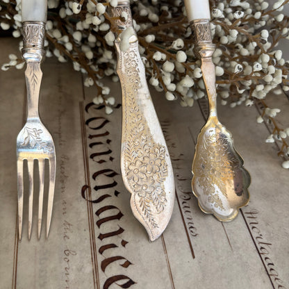Decorative silver fork, spoon, and knife with intricate patterns on a textured surface with white flowers in the background.