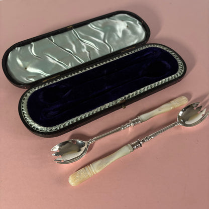 Silver spoons with ivory handles on a pink surface next to an open case.