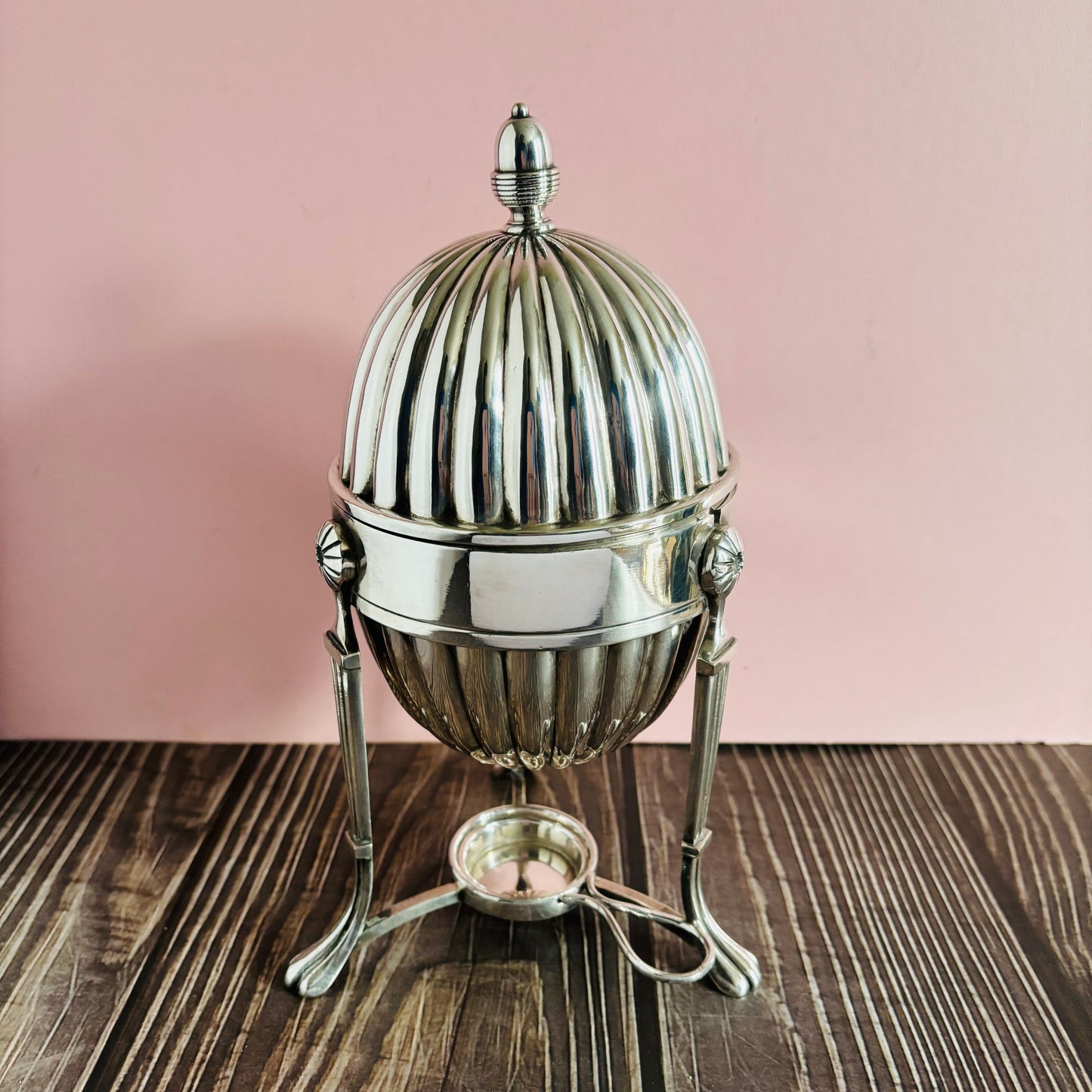 Silver egg-shaped container with a lid on a wooden surface against a pink background