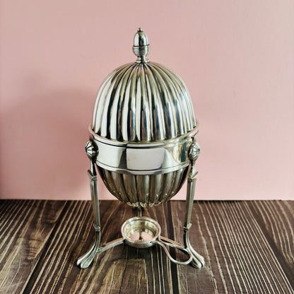 Silver egg-shaped container with a lid on a wooden surface against a pink background
