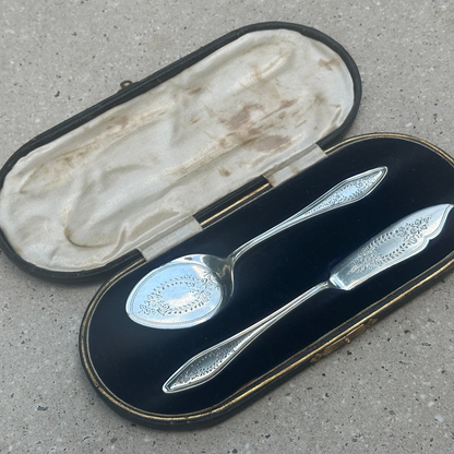 An antique pair of silver spoon and butter knife with ornate handles, presented in an open black velvet case with gold trim on a concrete surface.
