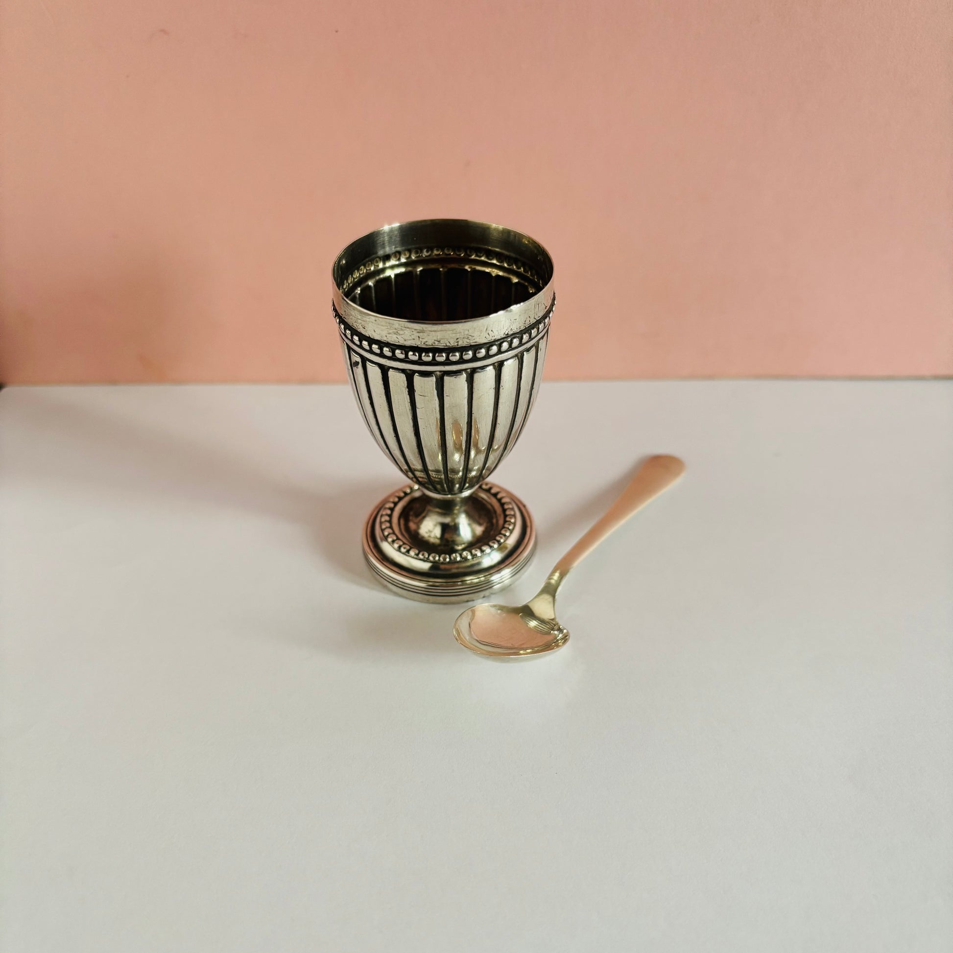 Silver egg cup with decorative patterns on a white surface against a pink background
