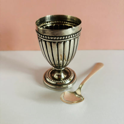 Silver goblet  shape egg cup and spoon on a white surface with a pink background