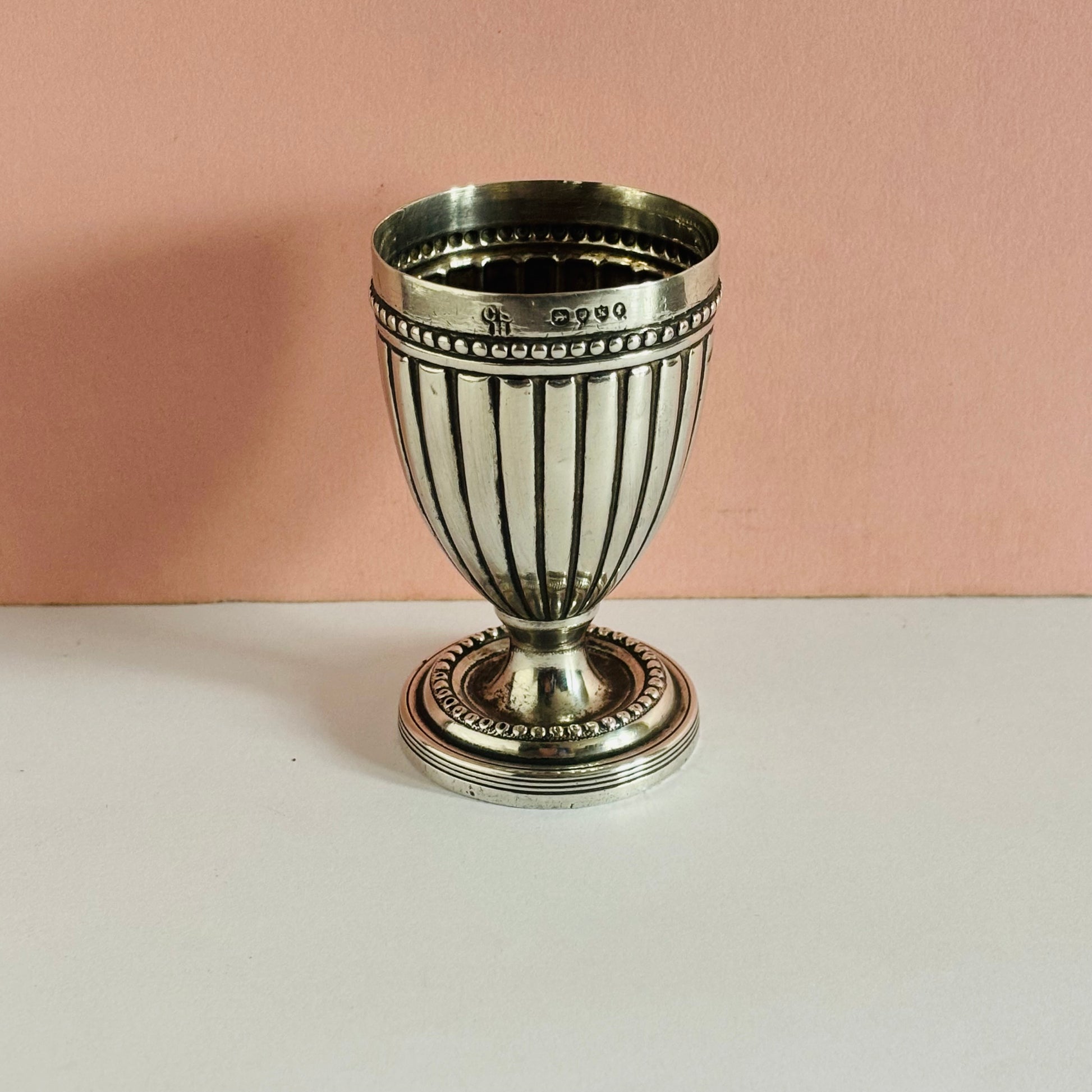 Silver goblet style eggcup with striped design on a white surface and pink background