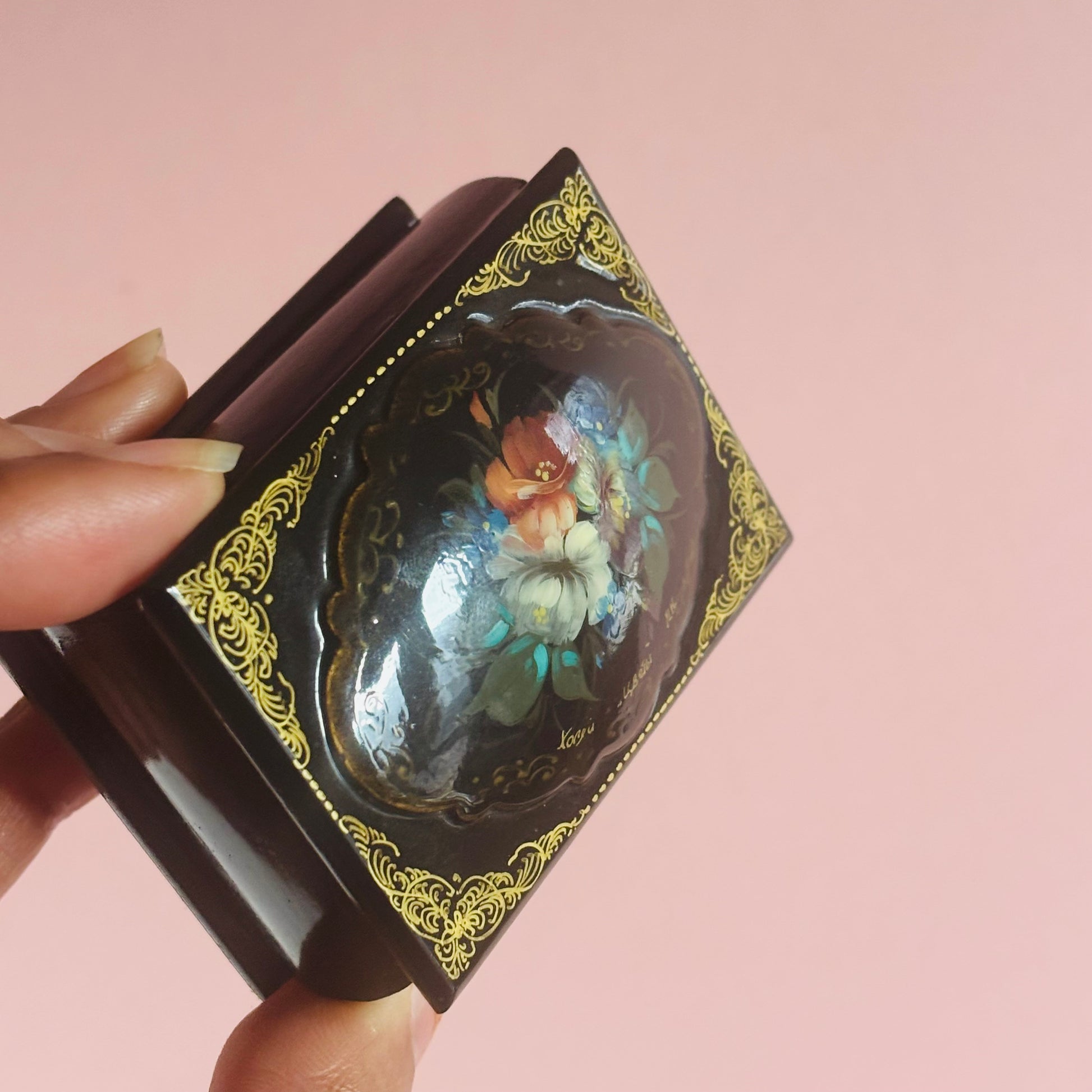 Vintage Paper Mache Lacquer Signed Small Russian Box with floral design held by a hand against a pink background