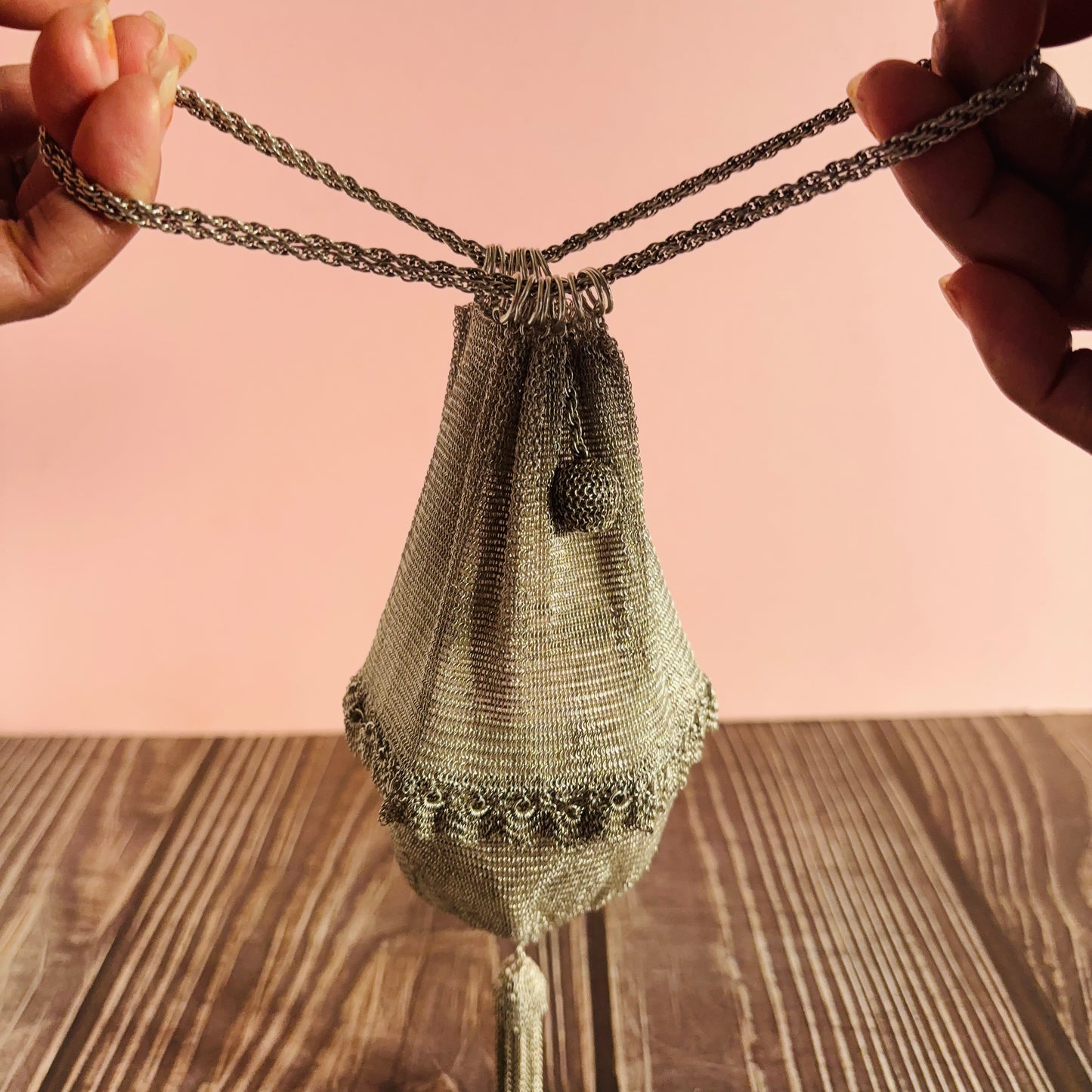 Handmade silver mesh bag with chain handle held by hands on a wooden surface with pink background