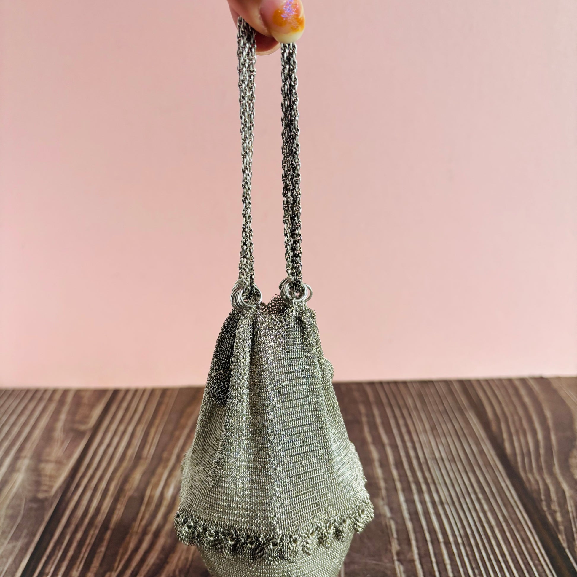 Silver mesh handbag with chain handle held by a hand on a wooden surface with a pink background