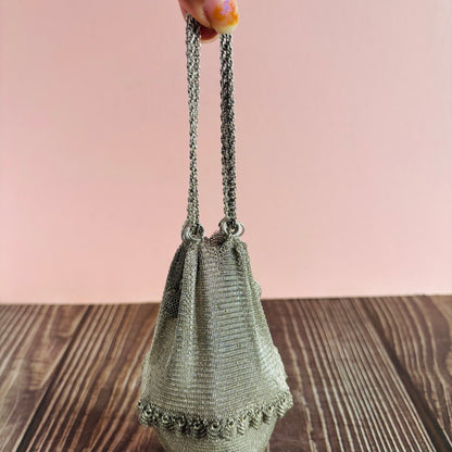 Silver mesh handbag with chain handle held by a hand on a wooden surface with a pink background