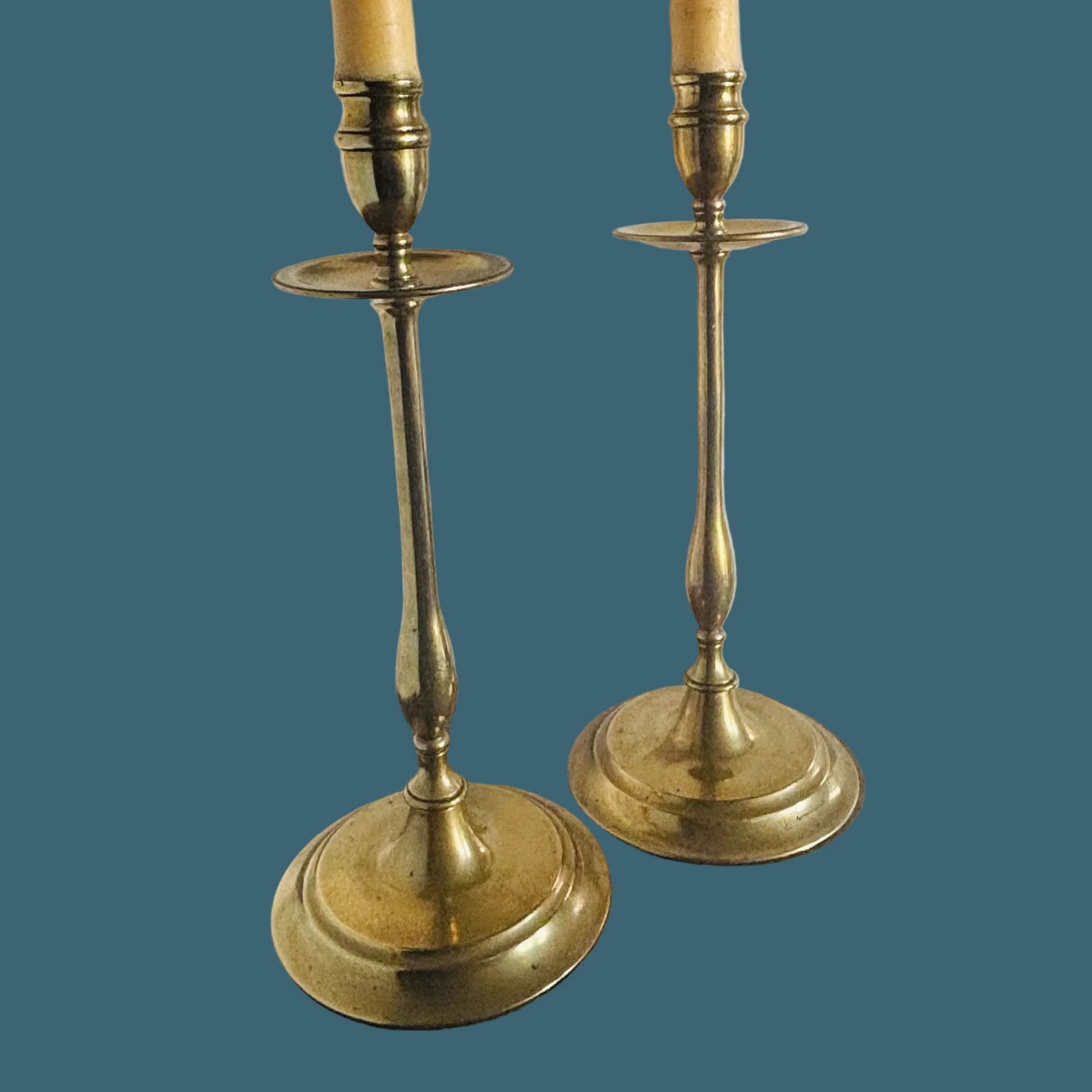 A pair of vintage brass candlestick holders with cream-colored candles, set against a solid dark teal background.