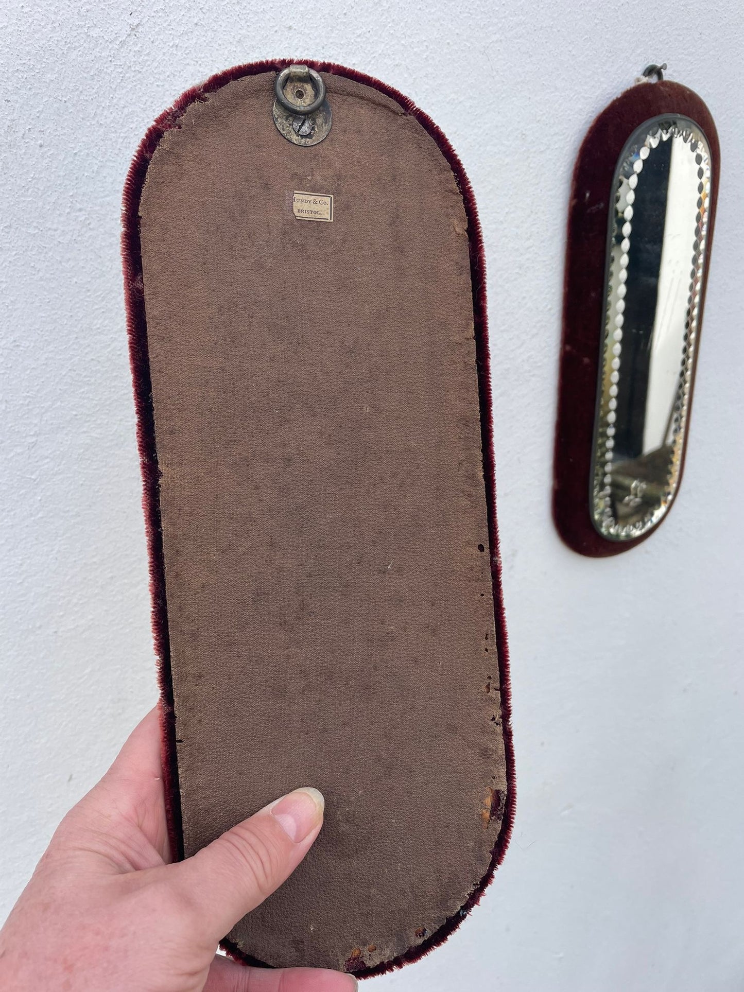 A hand holds a vintage oval wall mirror showing the back side with a brown velvet backing and dark red velvet edge. Another mirror is mounted on the white wall to the right.